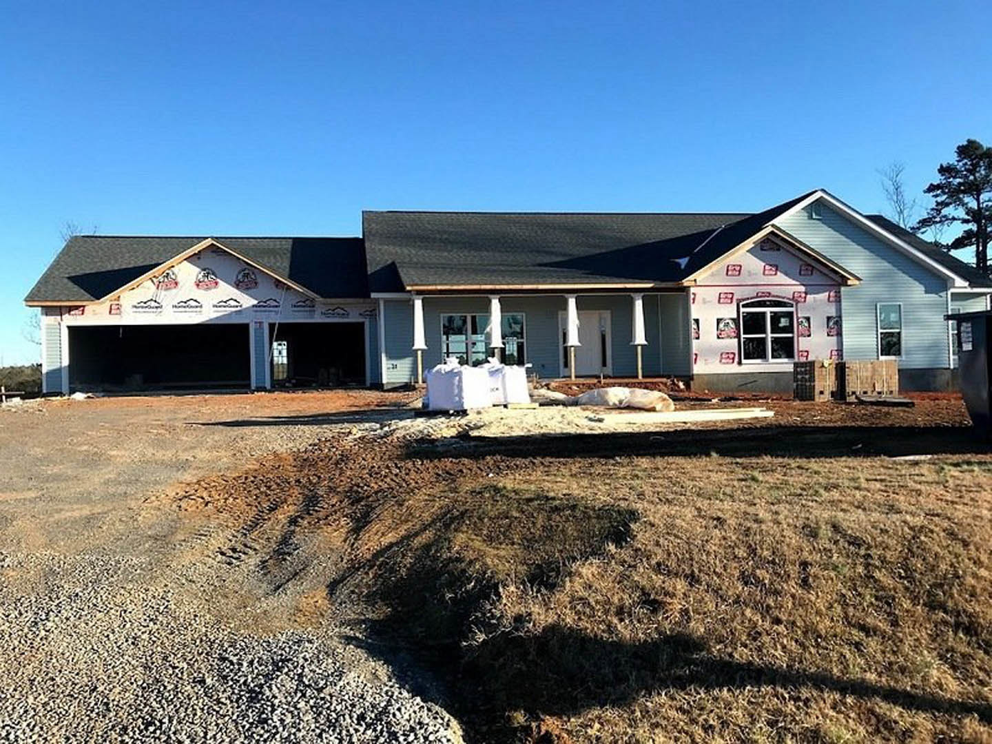 Framed house under construction with exposed pillars, dirt path leading to entrance, white siding, large leafy tree nearby, blue sky overhead, construction materials on pallet in