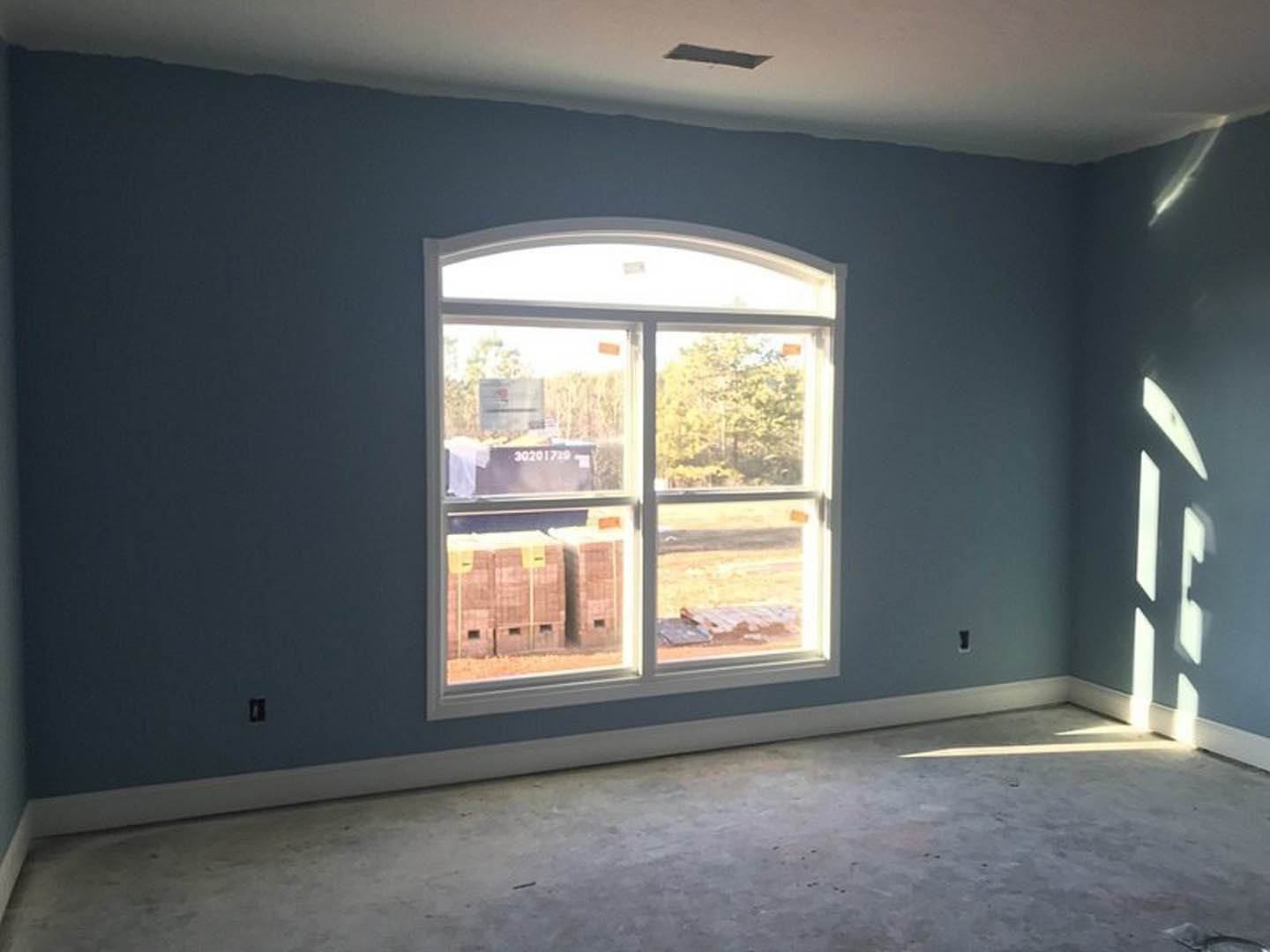 Concrete floor in a room with blue plaster walls, large window providing daylight, and stacked moving boxes near the door.