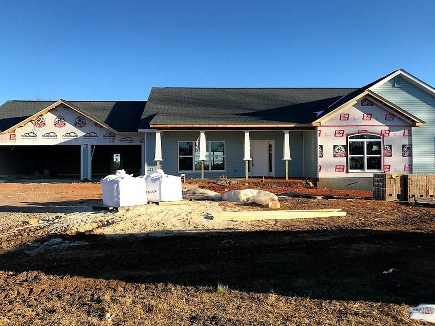 Partially built house with exposed framing, white columns, front door, construction materials and white bags scattered on ground, large window, hole in earth, blue sky overhead