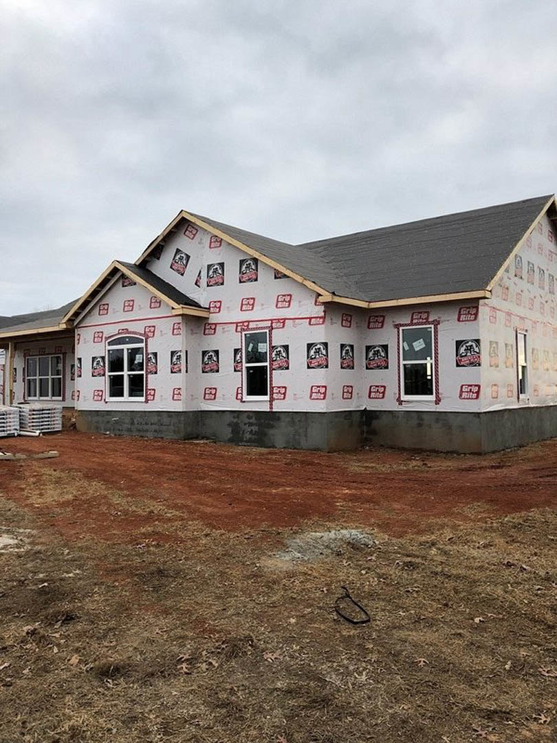 Partially built house with exposed sheathing secured by red and white adhesive tape, unfinished window, dirt and grass yard, cloudy sky overhead