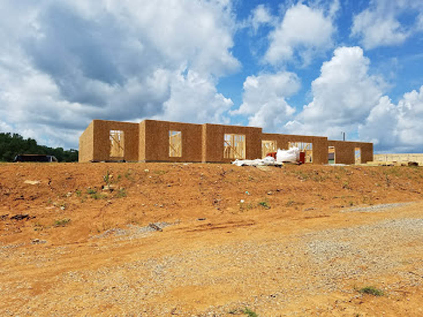 Framed custom home under construction on a hill with exposed wood, large window opening, dirt road in foreground, and cloudy sky overhead