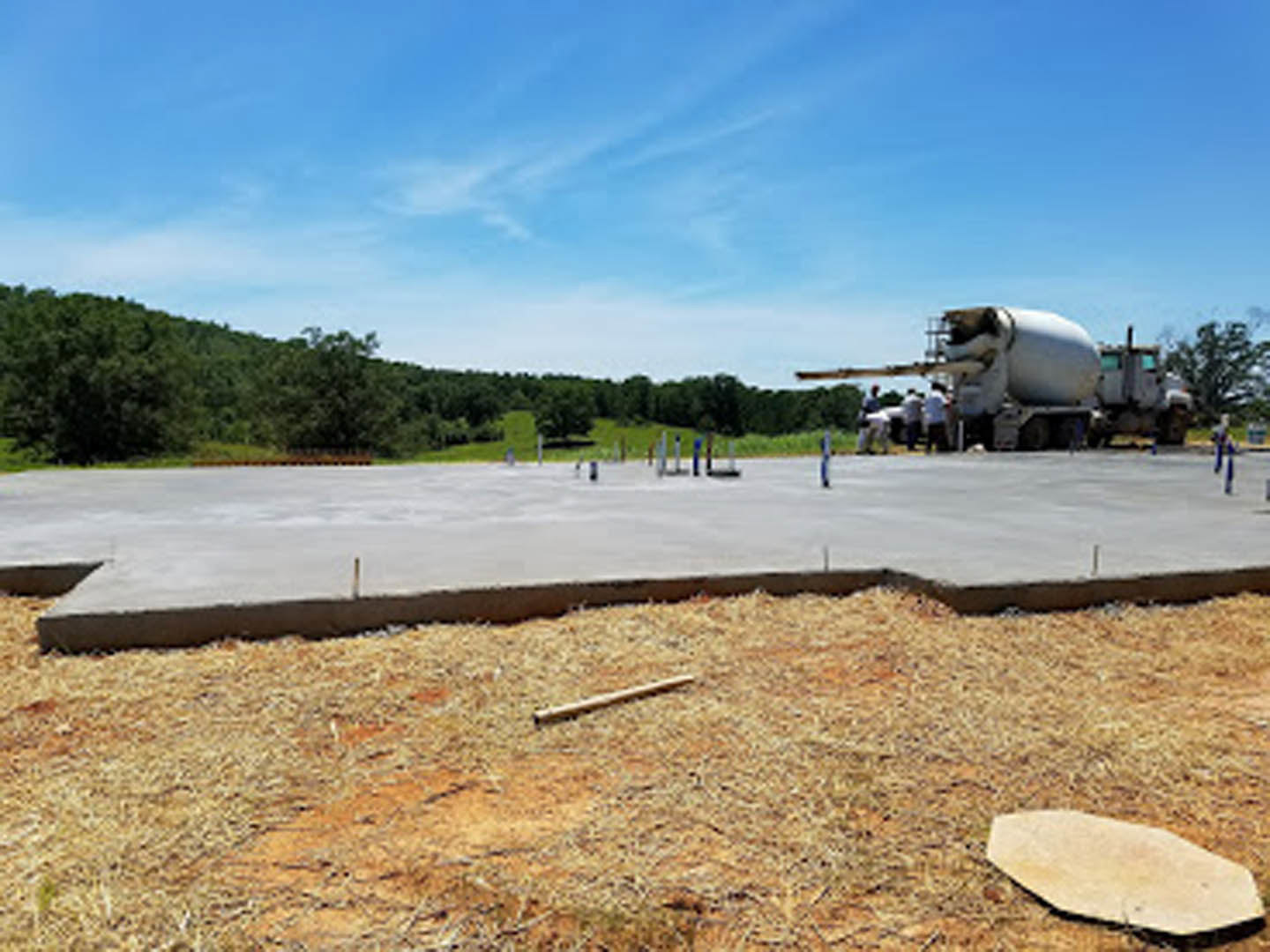 Concrete truck parked on dirt lot with several people standing on top, surrounded by scattered wood planks and a white oval container; grassy area and trees in background under