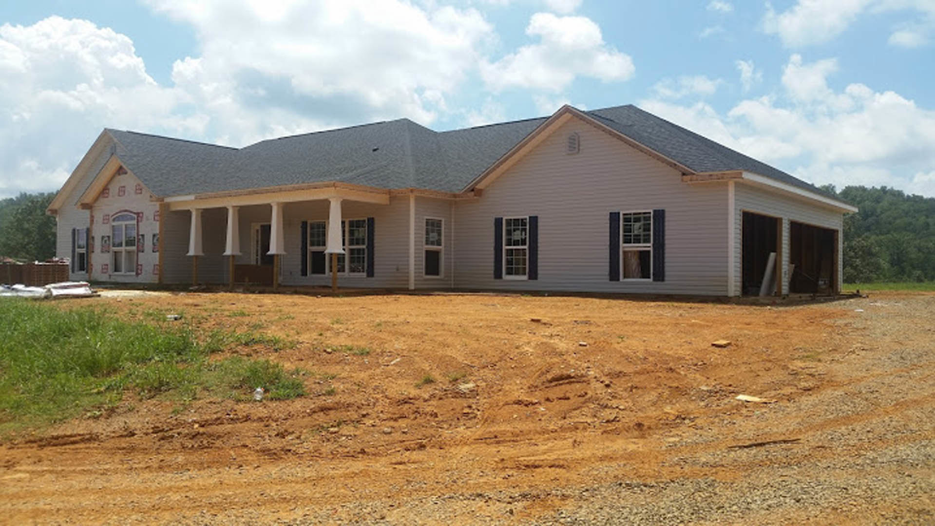 Two-story house with gray roof, white multi-pane windows, and dirt yard bordered by a metal fence and gravel road under a cloudy sky