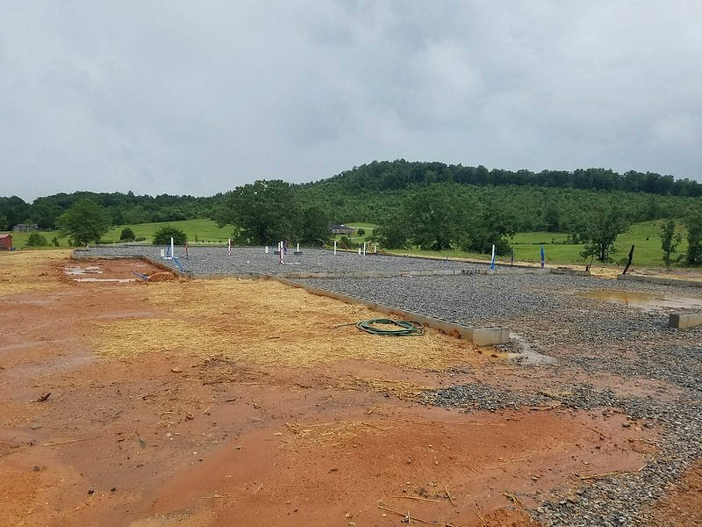 Dirt and gravel construction site bordered by trees and a hill, cloudy sky overhead, hose lying on ground, unfinished stonework visible