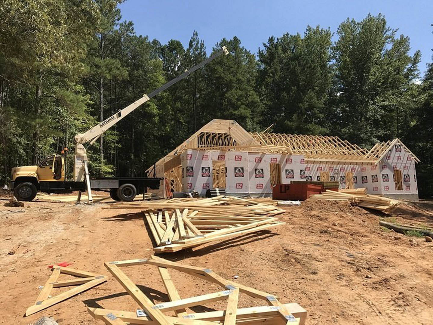 Wood-framed house under construction with exposed beams, crane and truck on site, pile of lumber and ladder nearby, surrounded by trees and open sky
