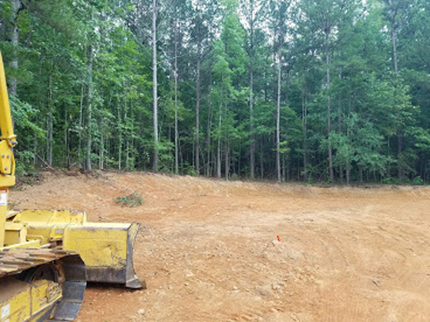 Yellow bulldozer parked on a dirt clearing surrounded by tall trees, forested background, exposed soil and scattered construction materials