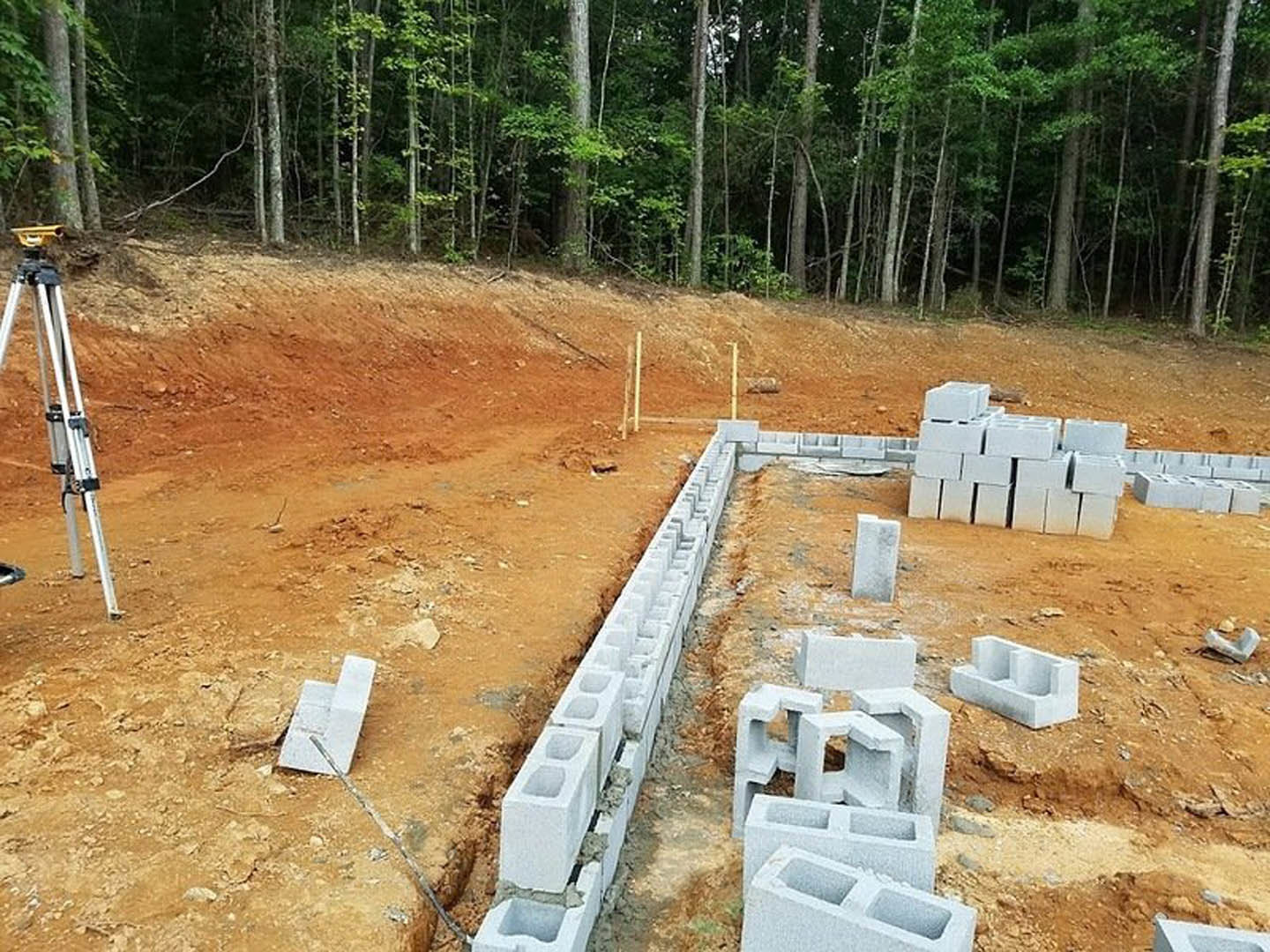 Partially built concrete block wall on construction site, surrounded by soil and scattered blocks, with trees in background and a metal ladder leaning nearby