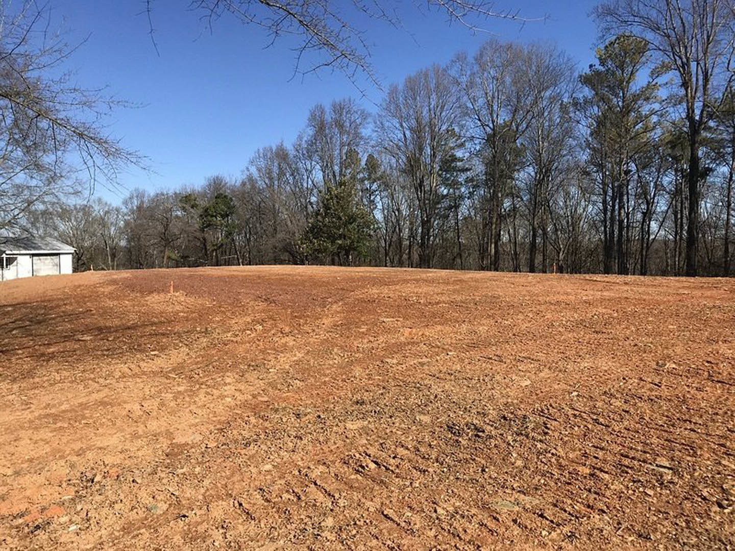 White custom home with garage door set on dirt field, surrounded by leafless and leafy trees under clear blue sky