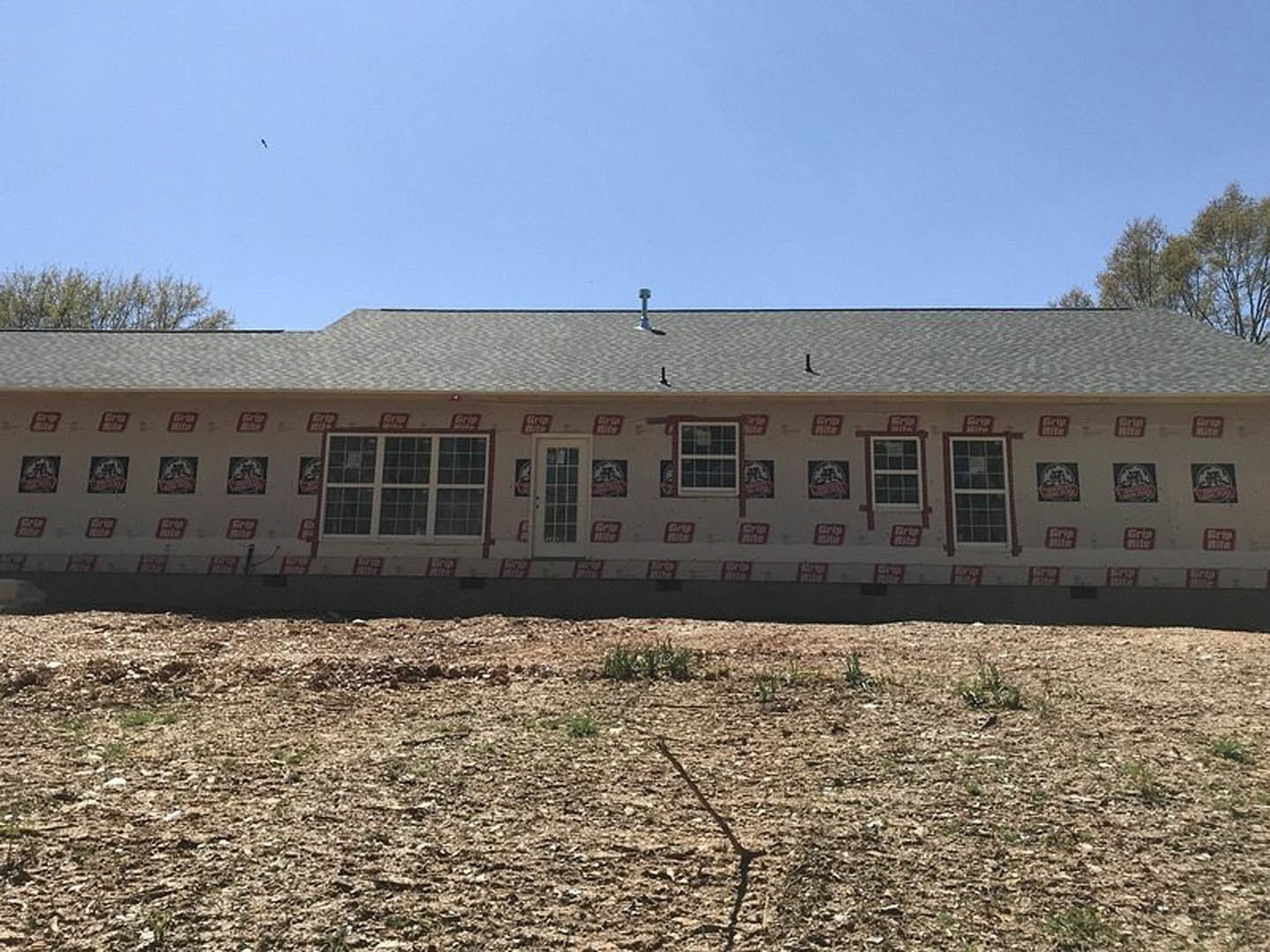 Two-story house with white siding and multiple windows, surrounded by a patchy dirt yard with sparse grass, trees in the background, and signs attached to the exterior wall.