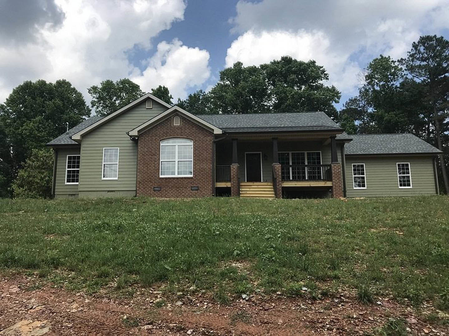 Red brick house with white-framed windows, covered front porch, black door, and stairs leading to a green lawn, surrounded by mature trees under a partly cloudy sky