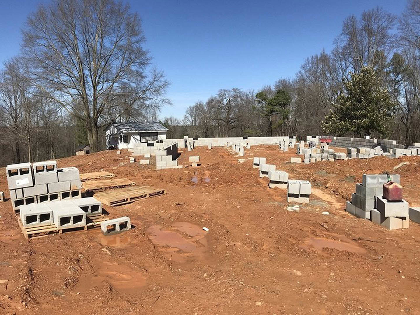 Cement blocks and pallets scattered across a residential construction site, partially built house with roof and window, large tree with white flowers, blue sky overhead