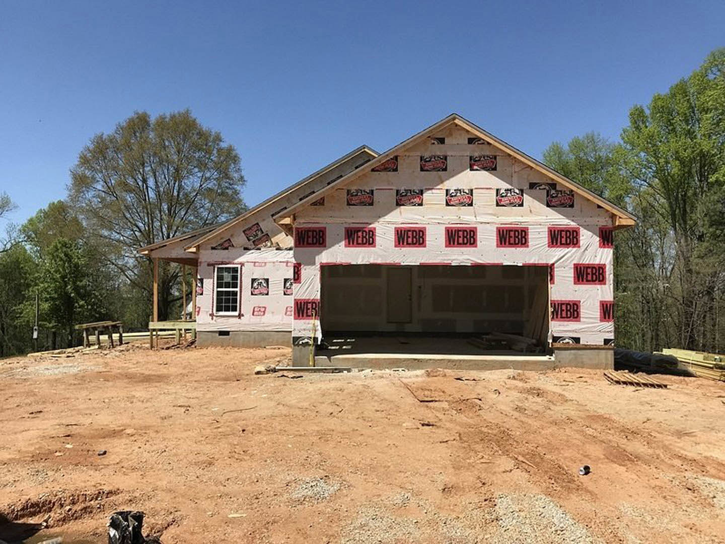 Partially built house with exposed framing, white window trim, red and white construction signs on exterior, dirt lot in foreground, tree and blue sky in background