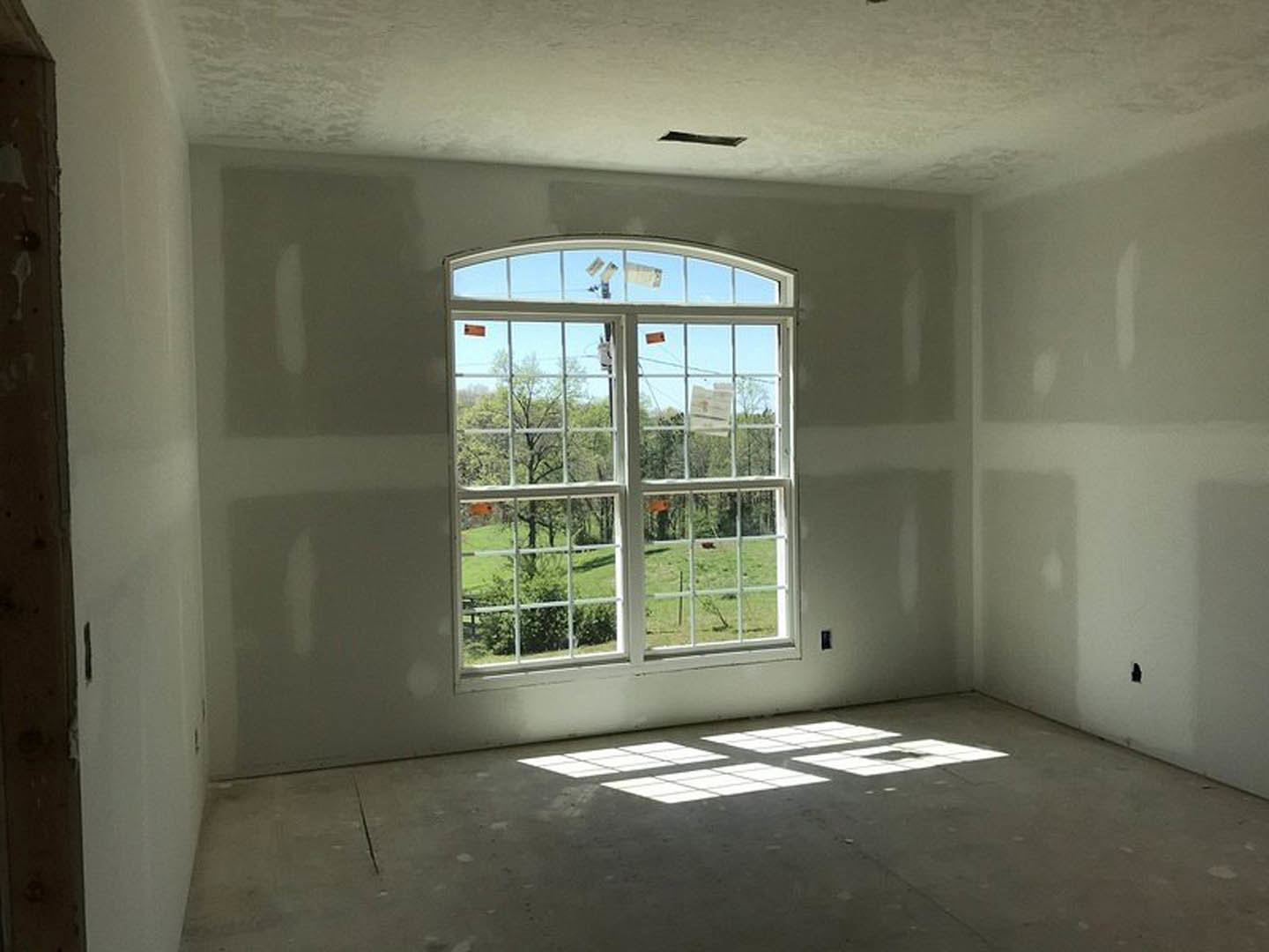 Sunlit room with smooth plaster walls, large window framing a grassy field and trees, natural light illuminating neutral finishes.