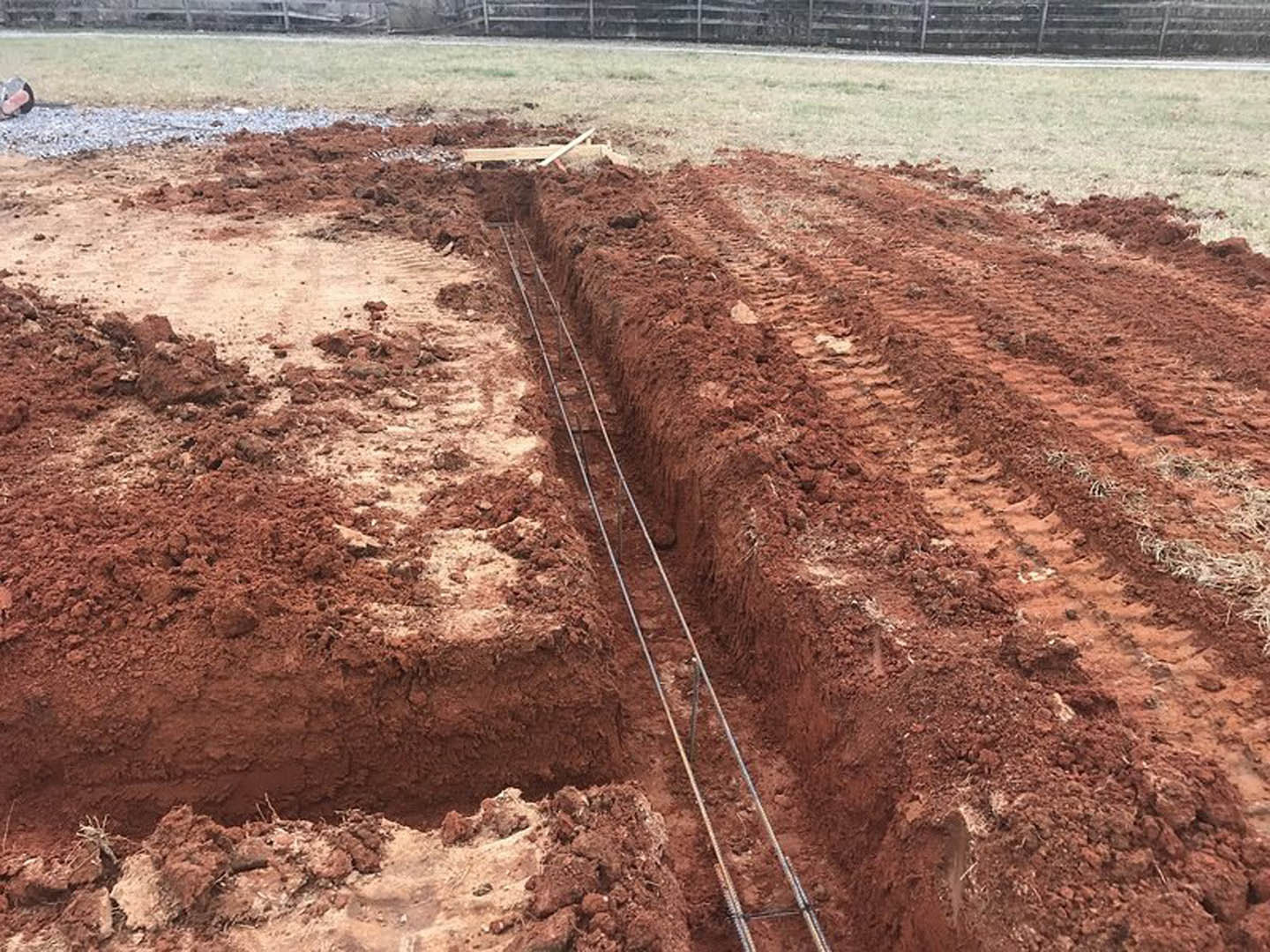 Wide trench with tire tracks in loose dirt, bordered by patches of grass and soil, construction site for home foundation