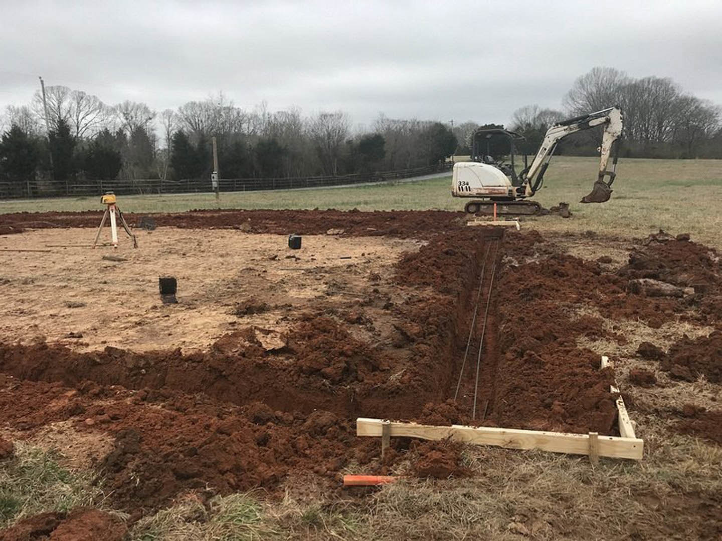 Bulldozer parked beside a freshly dug trench with exposed soil, wooden board placed in the ground, man standing nearby, cloudy sky and forested backdrop