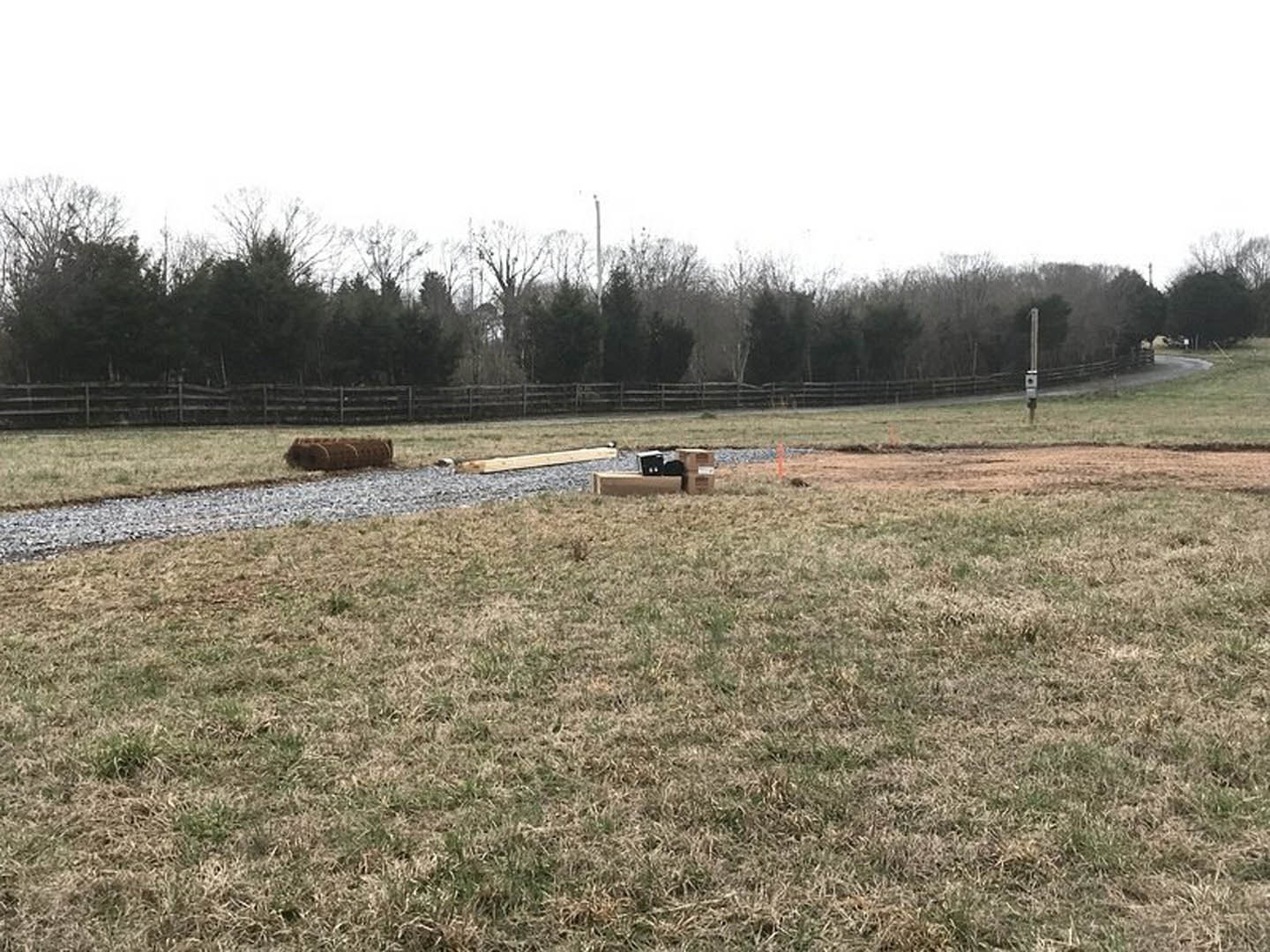 Grassy field bordered by wooden fences, leafless trees scattered throughout, gravel path leading past hay bales and black utility box