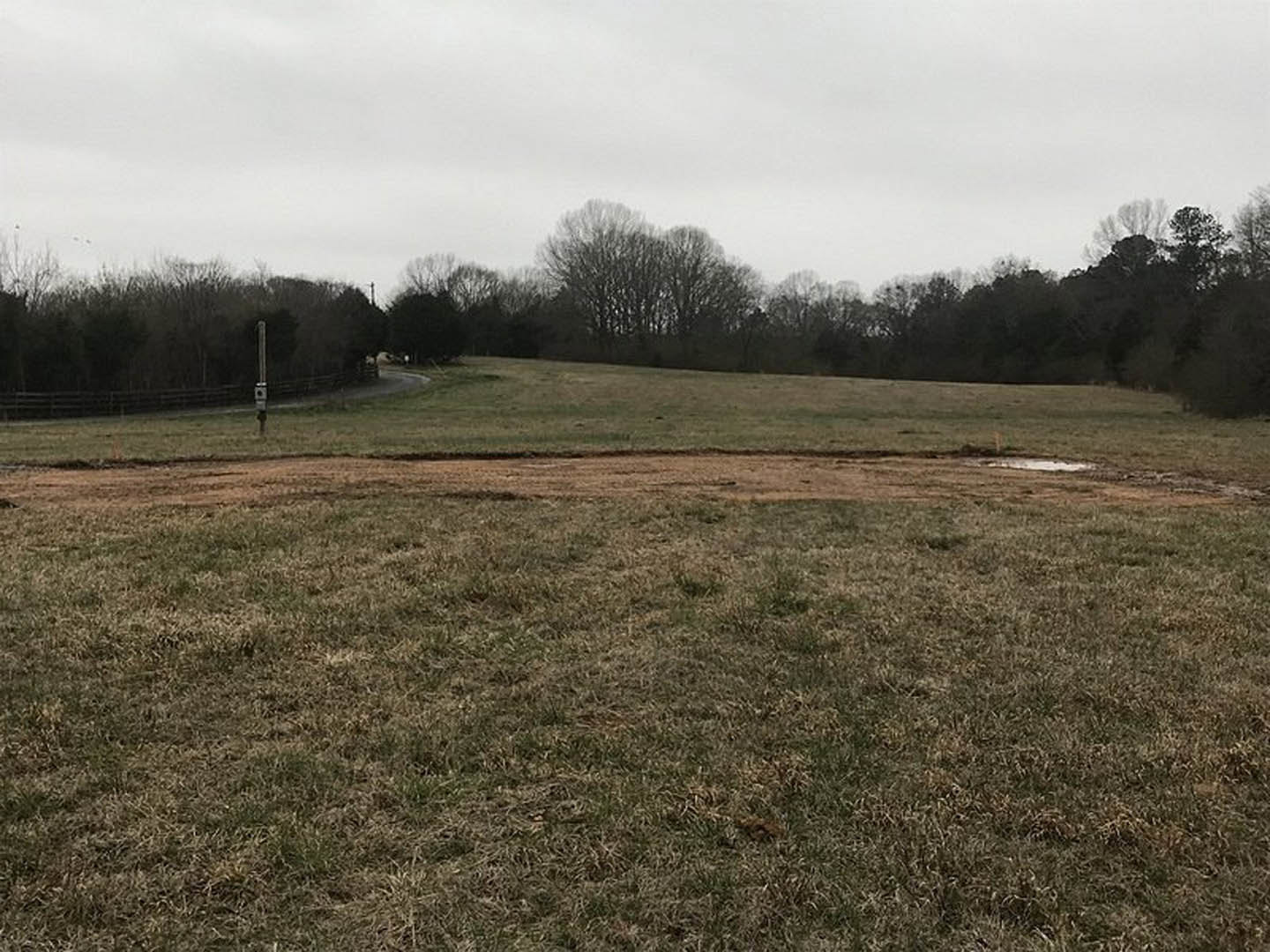 Grassy field with a dirt road, scattered trees in the background, wooden fence along the edge, partly cloudy sky overhead