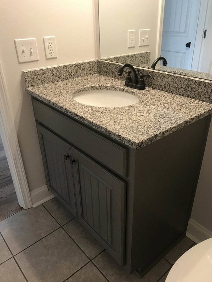 White quartz countertop with black specks, undermount sink, chrome faucet, rectangular mirror, white tile backsplash, double light switch, wall outlet, and gray cabinetry in a