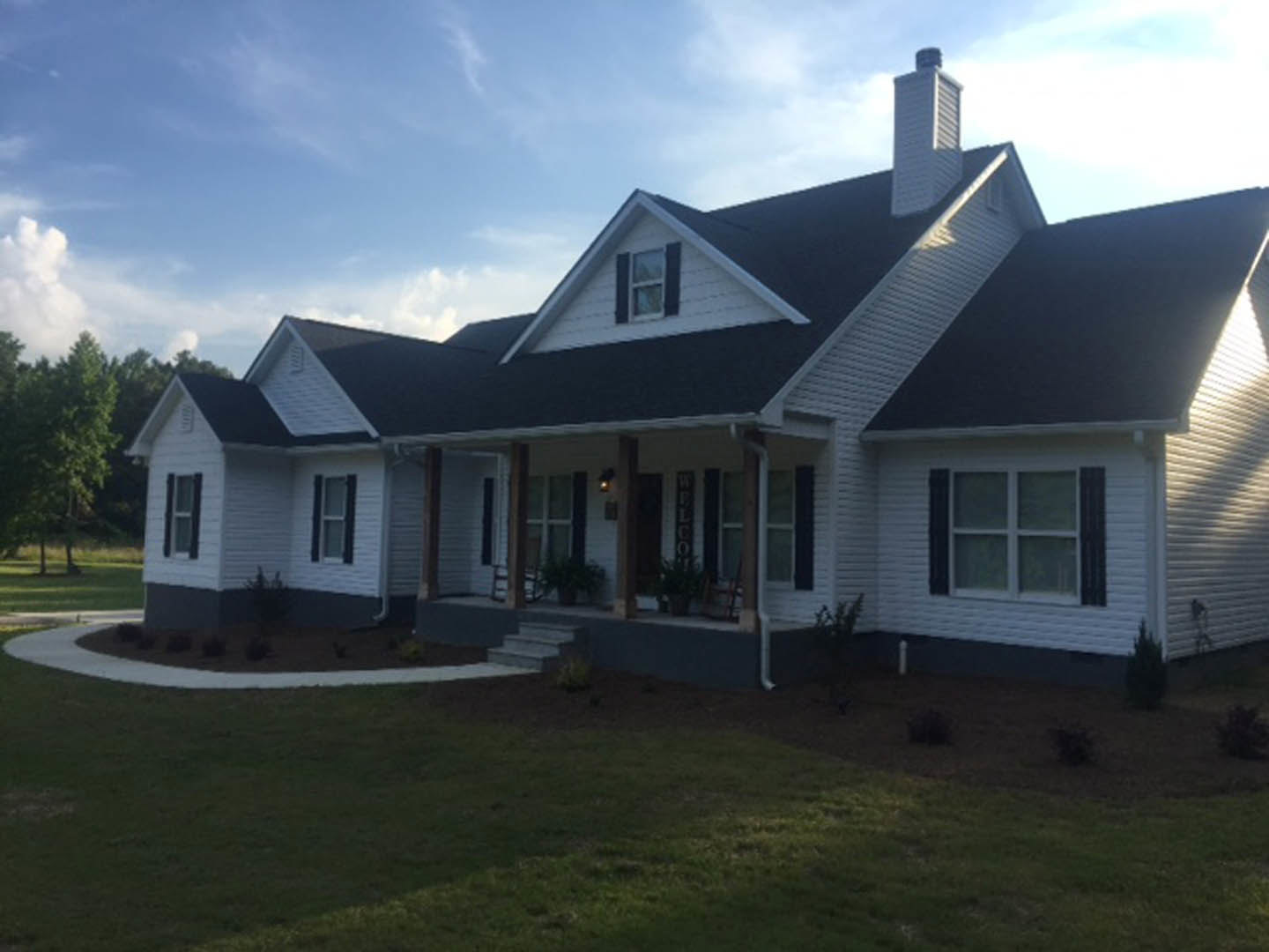 Two-story house with white siding, black shutters, covered front porch, manicured lawn, and blue sky with scattered clouds