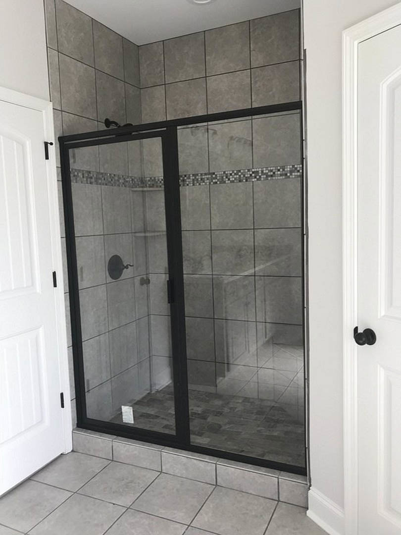 Frameless glass shower enclosure with chrome hardware, adjacent white door featuring matte black handle, light gray tile flooring and walls