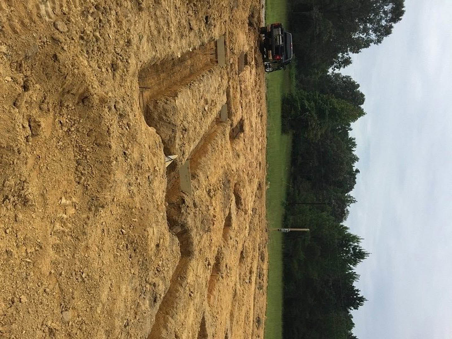 Brown dirt field with a pickup truck parked near a hill, surrounded by scattered trees, telephone pole, and cloudy sky overhead