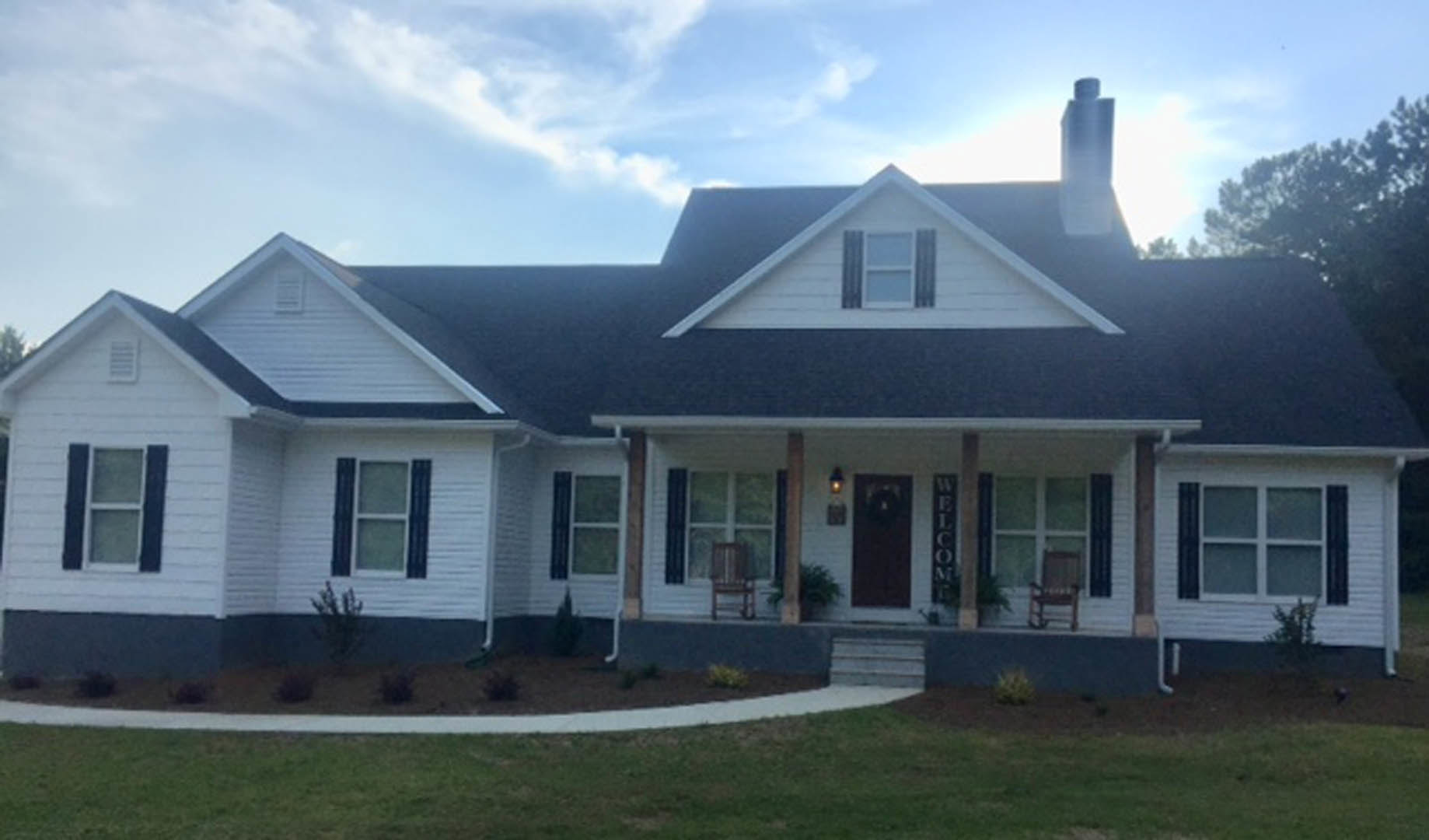 Two-story home with white siding, covered front porch, black shutters, manicured green lawn, and bright blue sky with scattered clouds