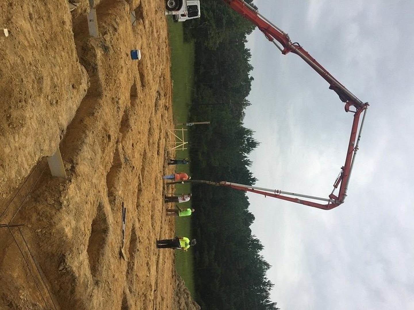 Construction site with group of people standing on dirt field, red crane lifting concrete beam, truck and metal rods visible, cloudy sky and trees in background