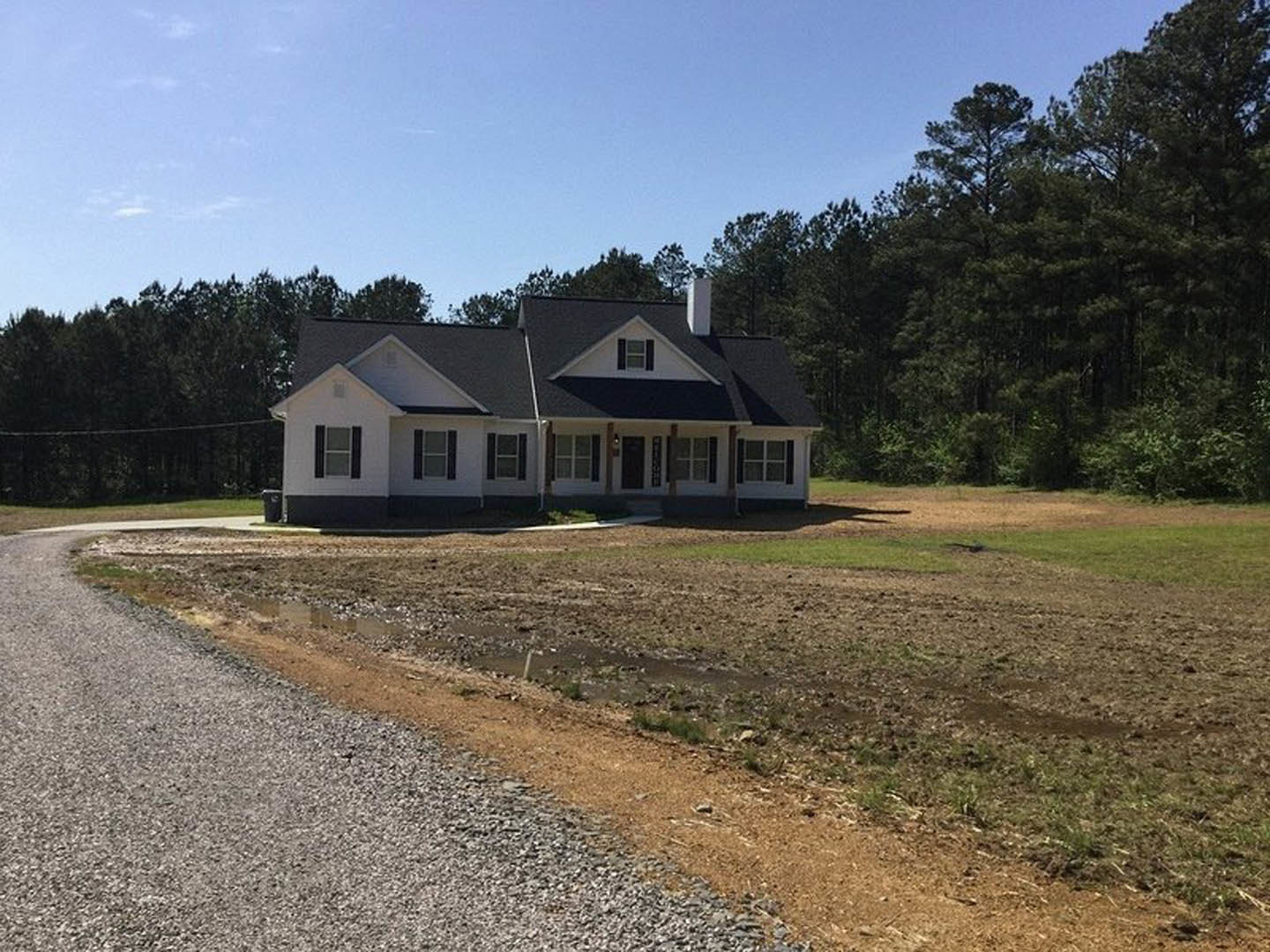Black-roofed cottage with white-framed windows, surrounded by trees and grass, dirt road running alongside the house under a cloudy sky