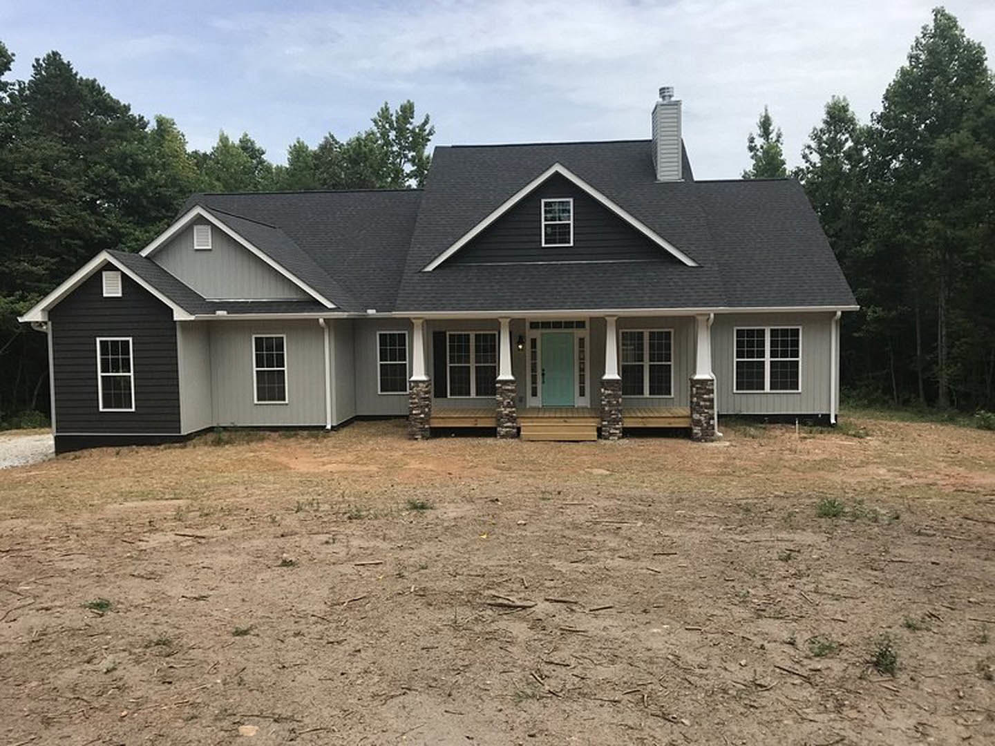 Two-story house with beige siding, blue front door, white-framed windows, covered porch, and dirt yard under partly cloudy sky