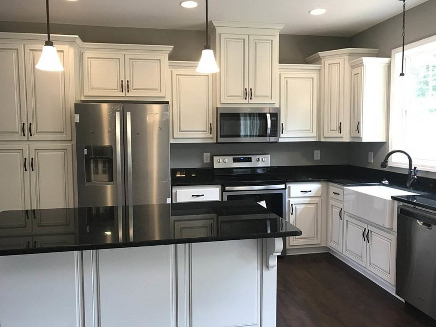 White kitchen cabinets with black countertops, stainless steel microwave, refrigerator, and stove, white window letting in natural light, smooth painted wall