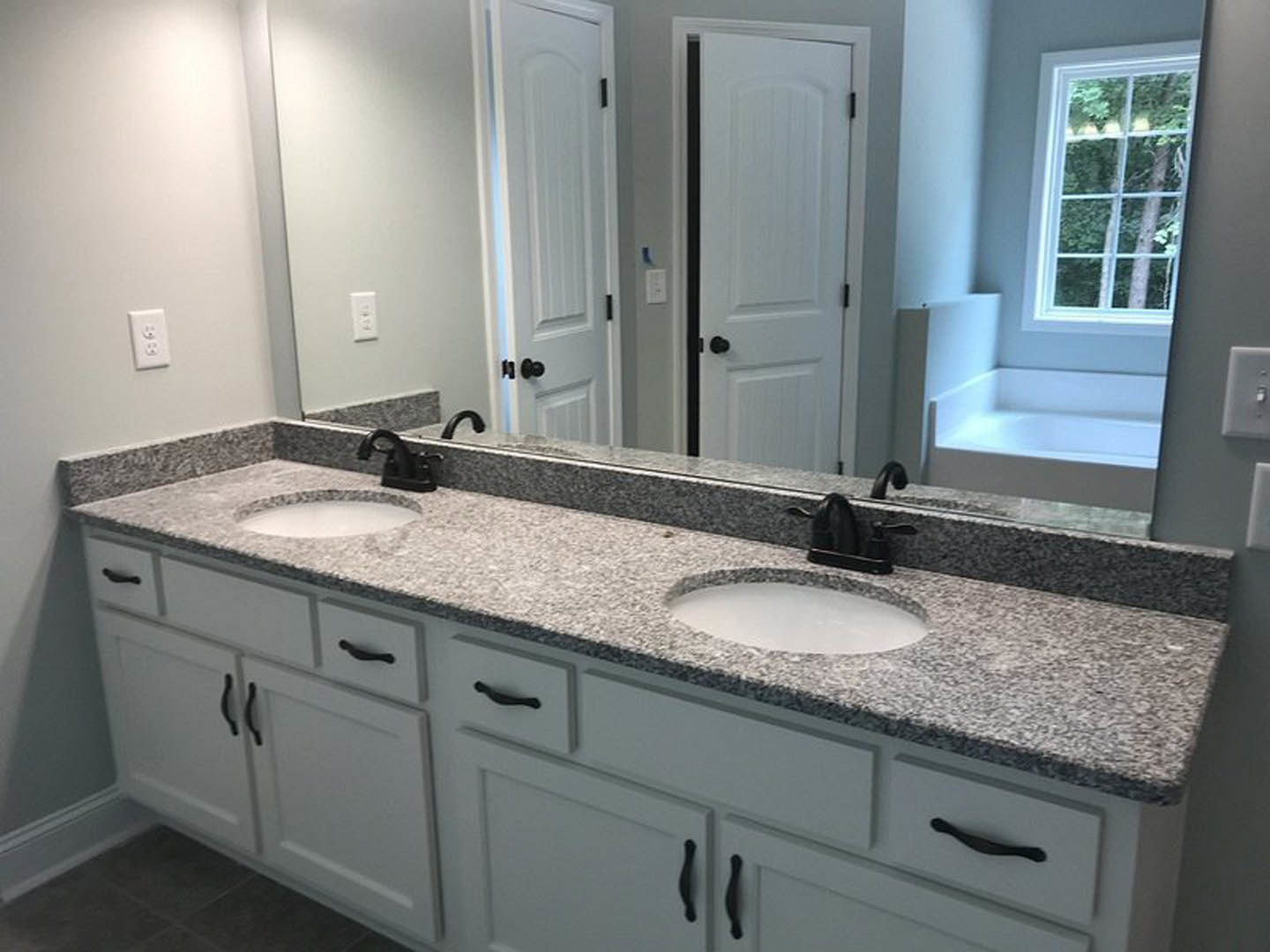 Bathroom with double vanity sinks, freestanding bathtub, white cabinetry, stone countertops, large multi-pane window, chrome faucets, and light-colored tile flooring.