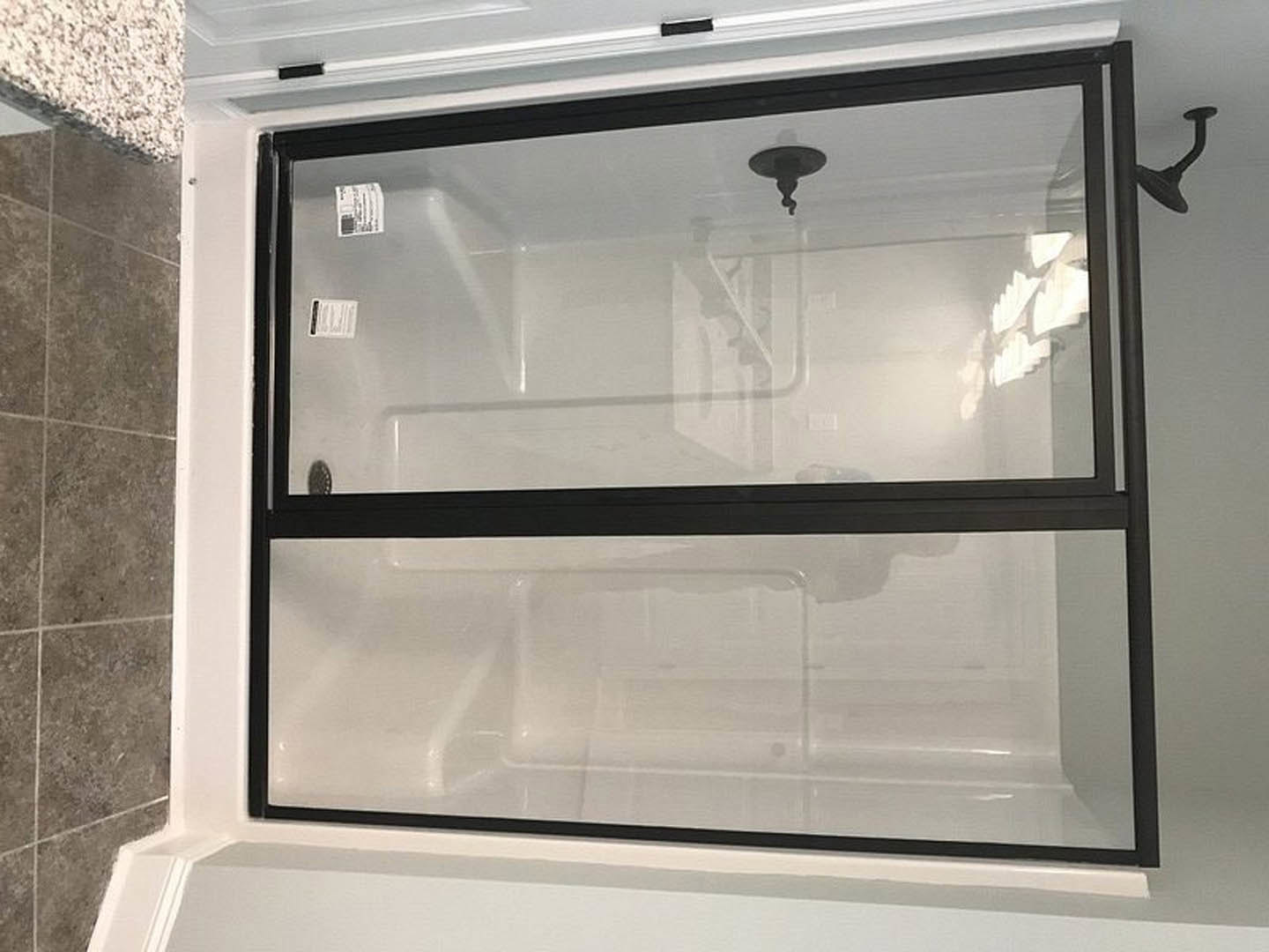 Frameless glass shower door with chrome handle in white tiled bathroom, gray floor tiles, partial view of window and wall mirror