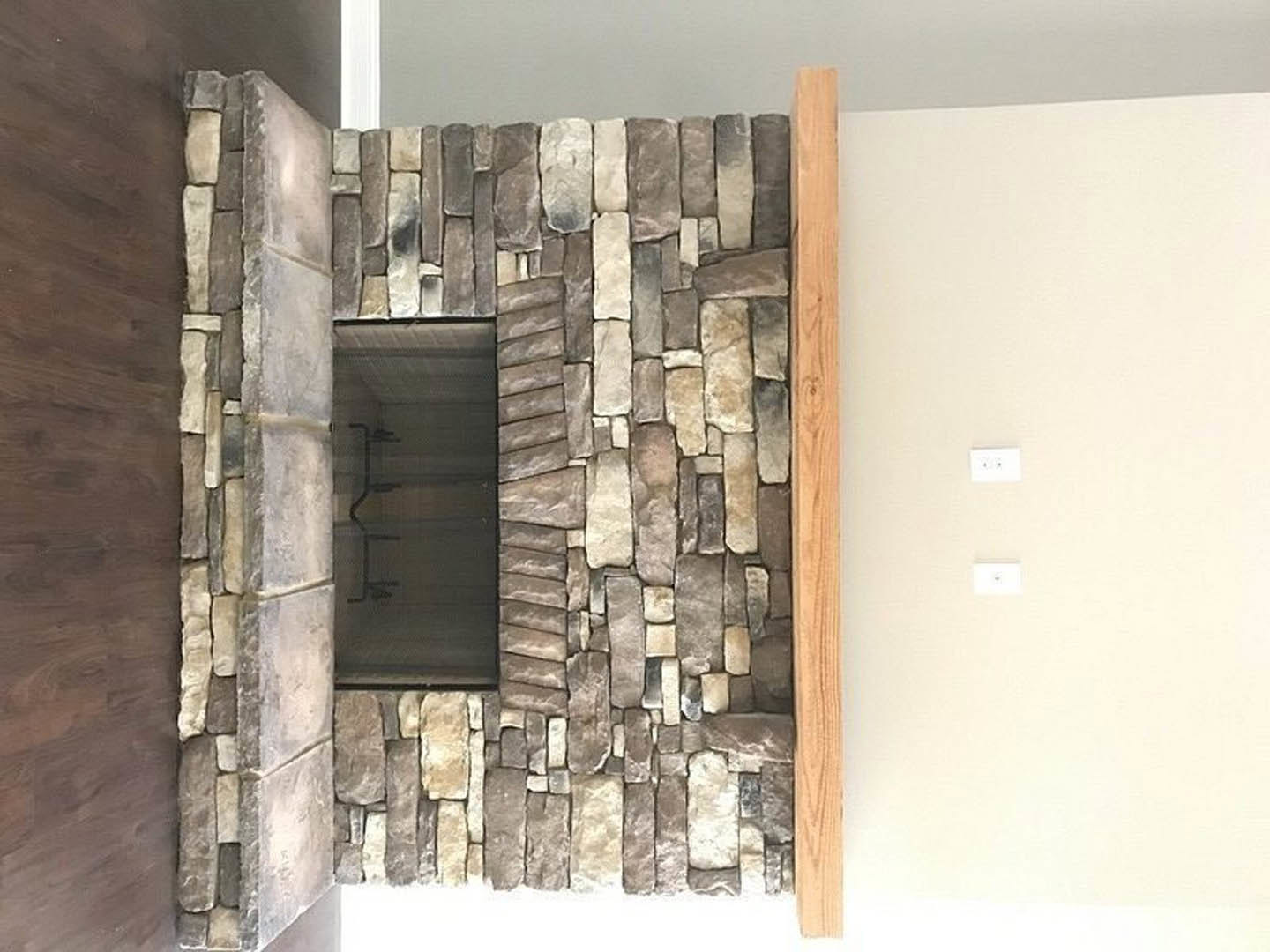 Stone fireplace with stacked stone surround and wooden mantel shelf, adjacent white plaster wall featuring a light switch and black and white logo artwork, window beside fireplace