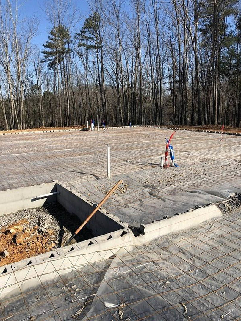Concrete foundation under construction with a shovel resting nearby, surrounded by dirt and scattered building materials, with a backdrop of tall trees and blue sky.