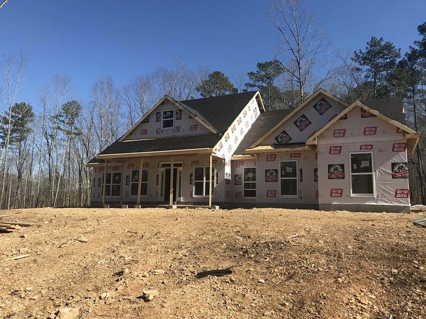 Partially built house with exposed wooden framing, installed roof and windows, surrounded by mature trees; construction materials scattered on bare ground.
