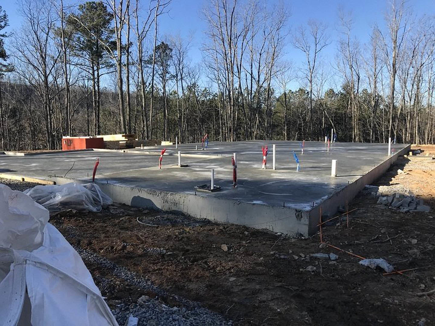 Concrete foundation slab with several metal poles installed at the center, surrounded by bare ground and trees in a wooded area, white tarp partially visible on the ground.
