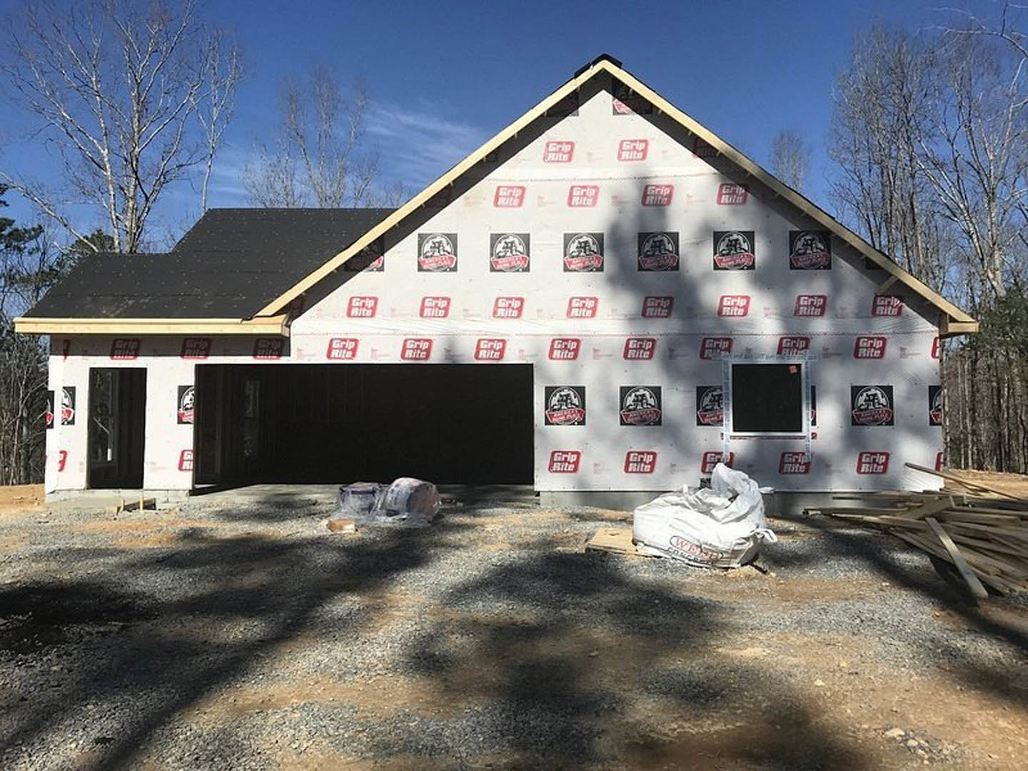 Partially built house with exposed metal framing, gravel and rocks covering the ground, white construction bag near the foundation, attached garage, surrounding trees, and clear