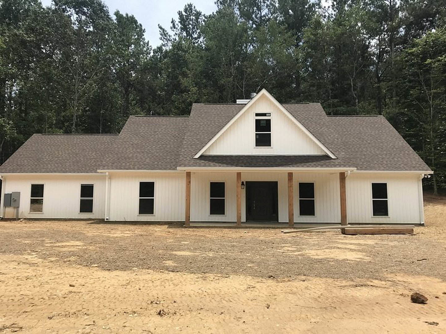 White siding house with brown shingle roof, black front door with exterior light, white-framed windows, dirt yard, distant building visible in background.