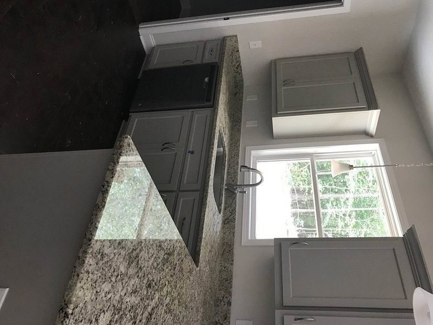Granite countertops with white cabinetry, grey cabinets featuring wire accents, white paneled wall with window, pendant lamp in front of window, stainless steel sink and appliances