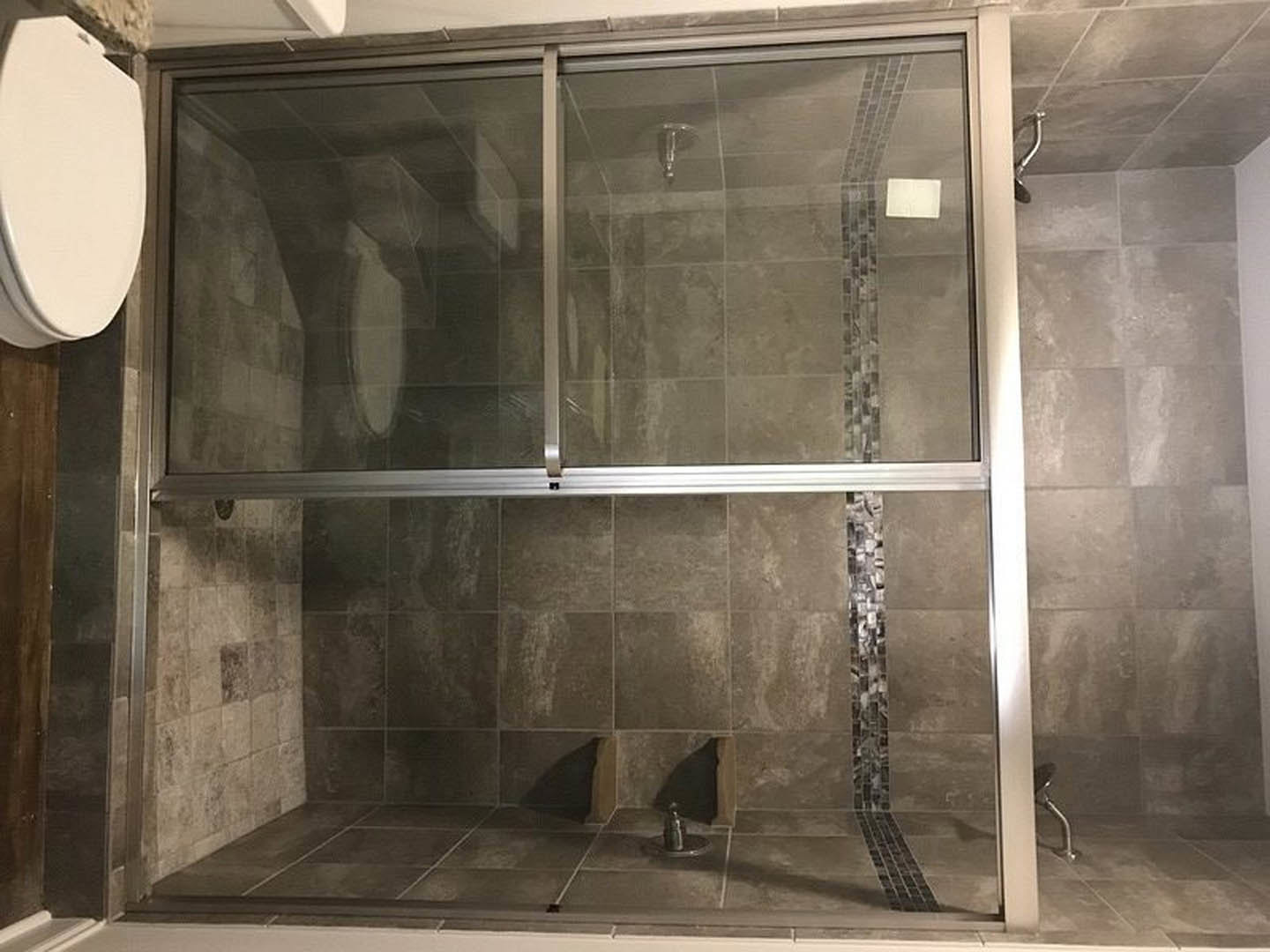 Walk-in shower with clear glass doors, gray tile walls, and a white toilet adjacent to a sliding glass door in a modern master bathroom.