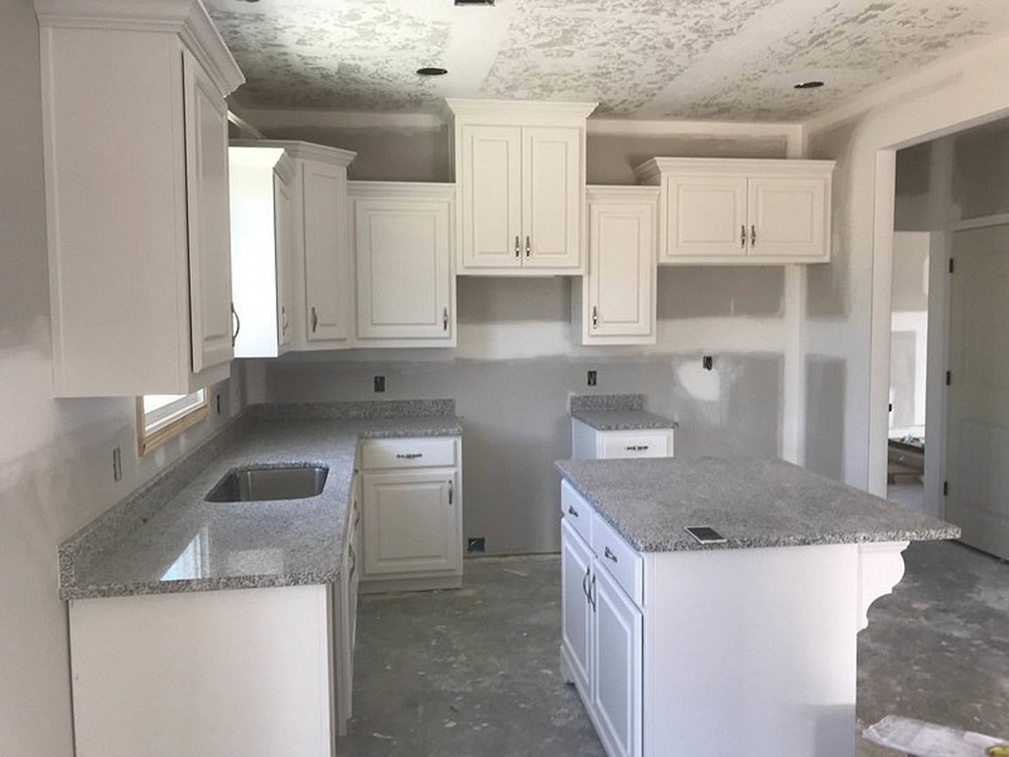 White kitchen with granite countertops, white cabinets with silver handles, kitchen island topped with granite, stainless steel sink, built-in appliances, and light walls.