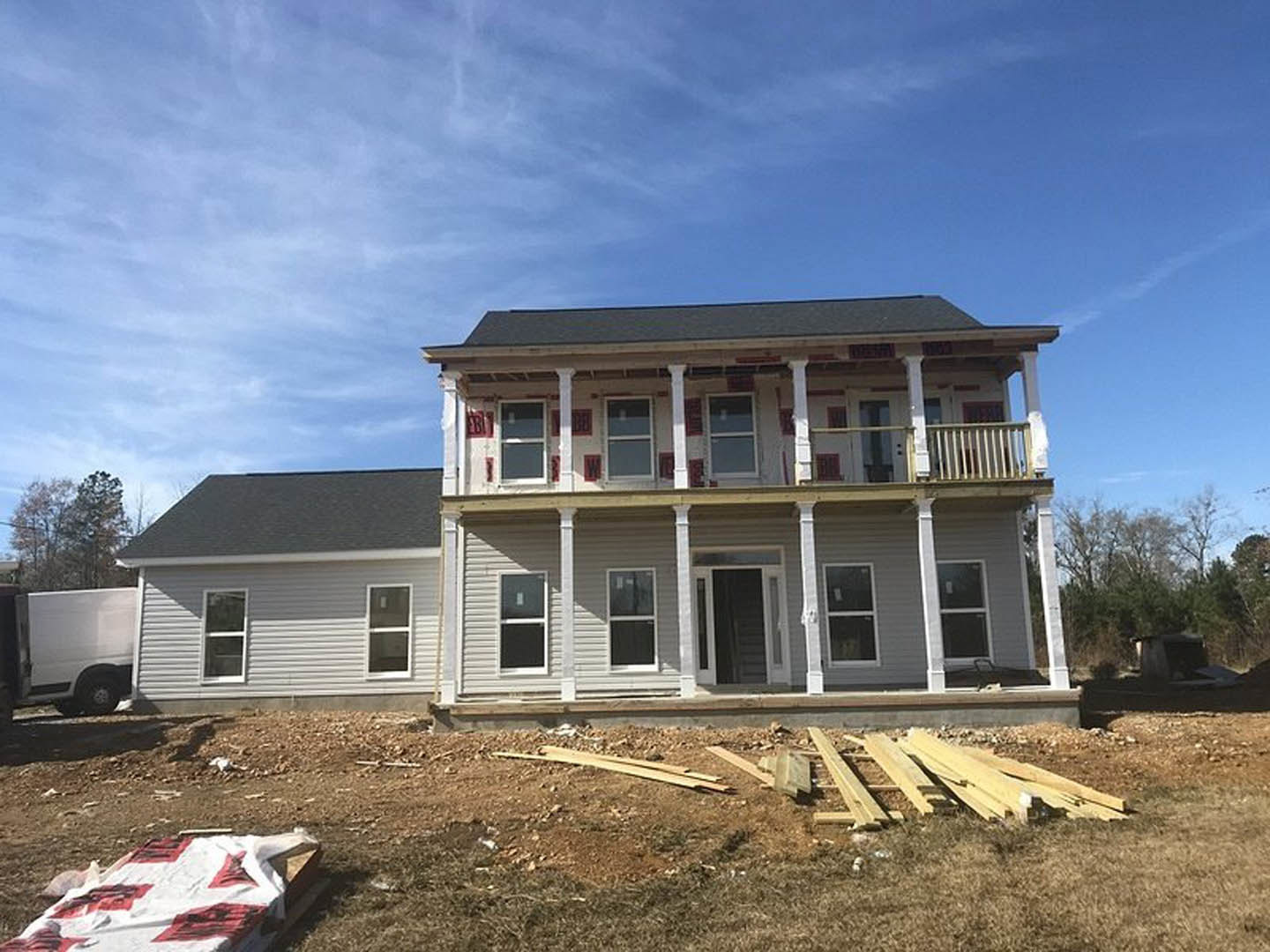 Partially built house with exposed framing, white pillars, piles of wood planks on dirt, white truck parked nearby, window with white frame, cloudy sky overhead