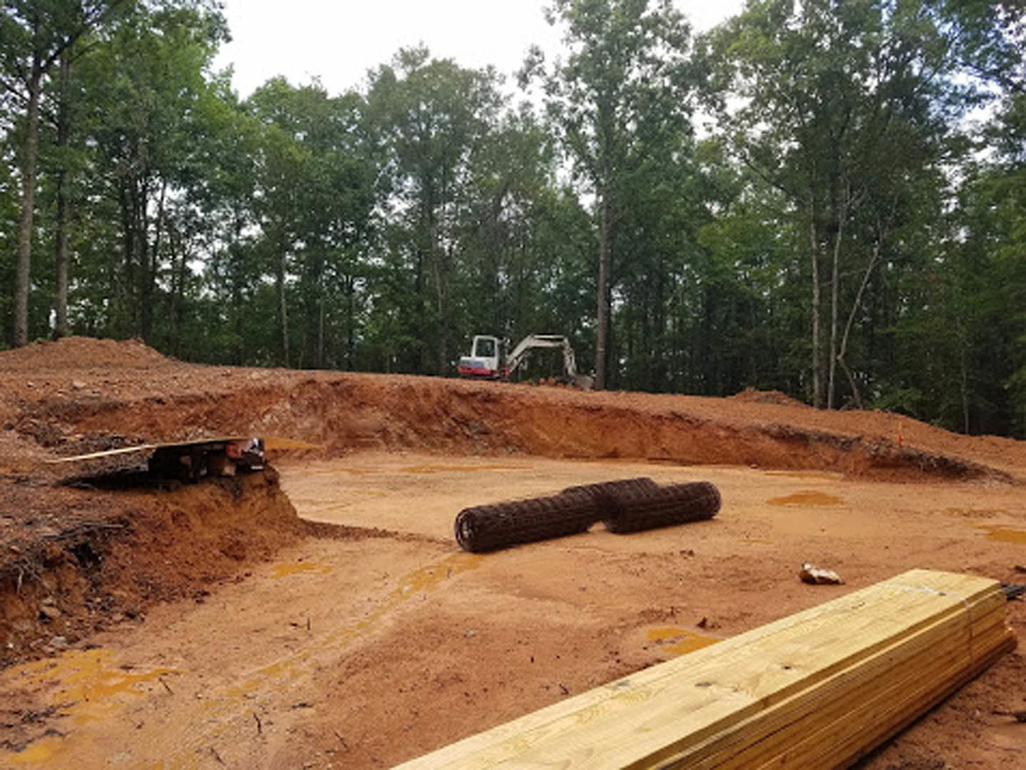 Large yellow loader parked on soil foundation surrounded by trees, stacks of lumber, and construction materials under a clear sky