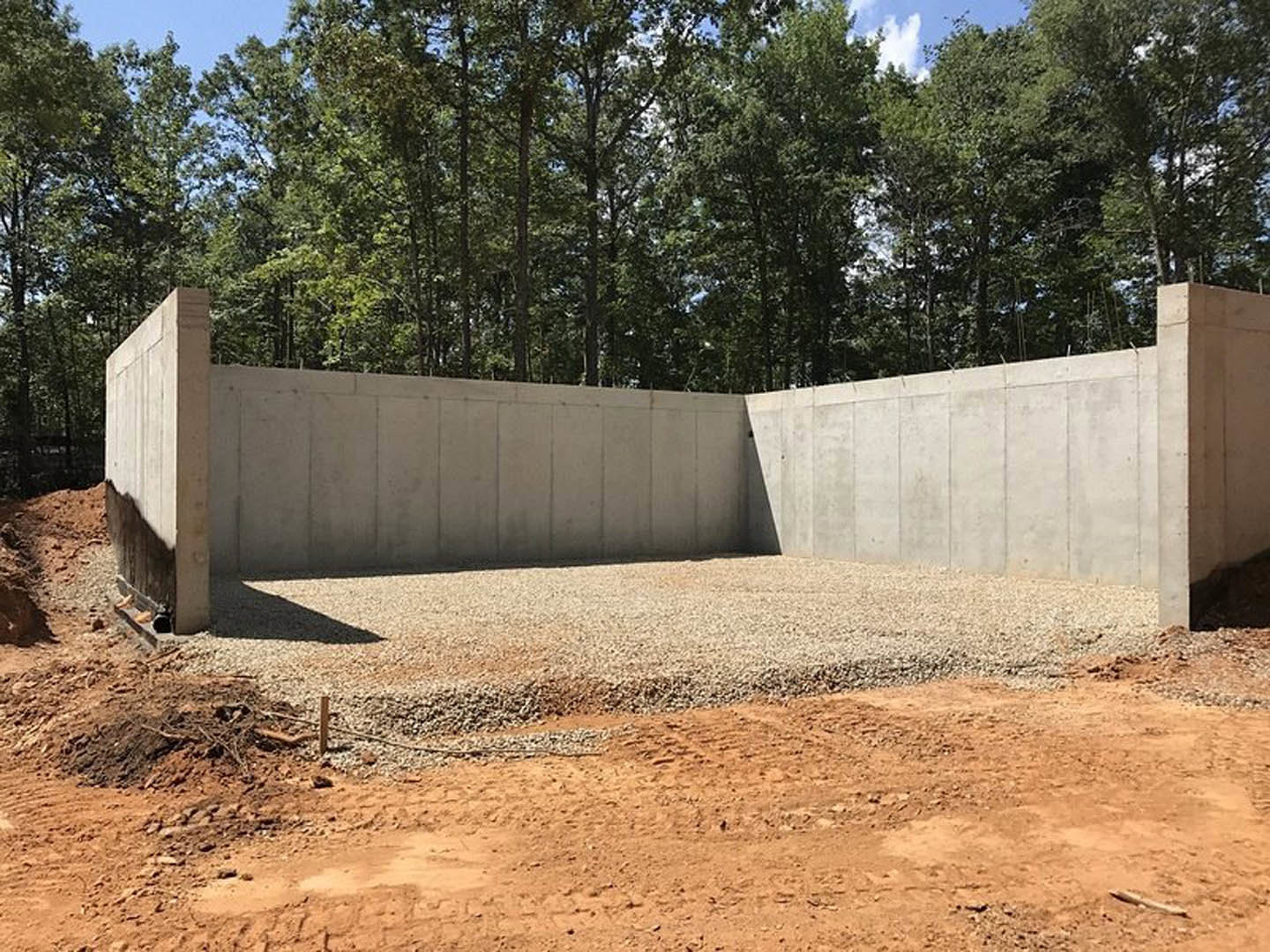 Concrete wall bordered by gravel, with trees and forested background, dirt ground with tire tracks, stone pillar, and shadow of a person's head visible in foreground