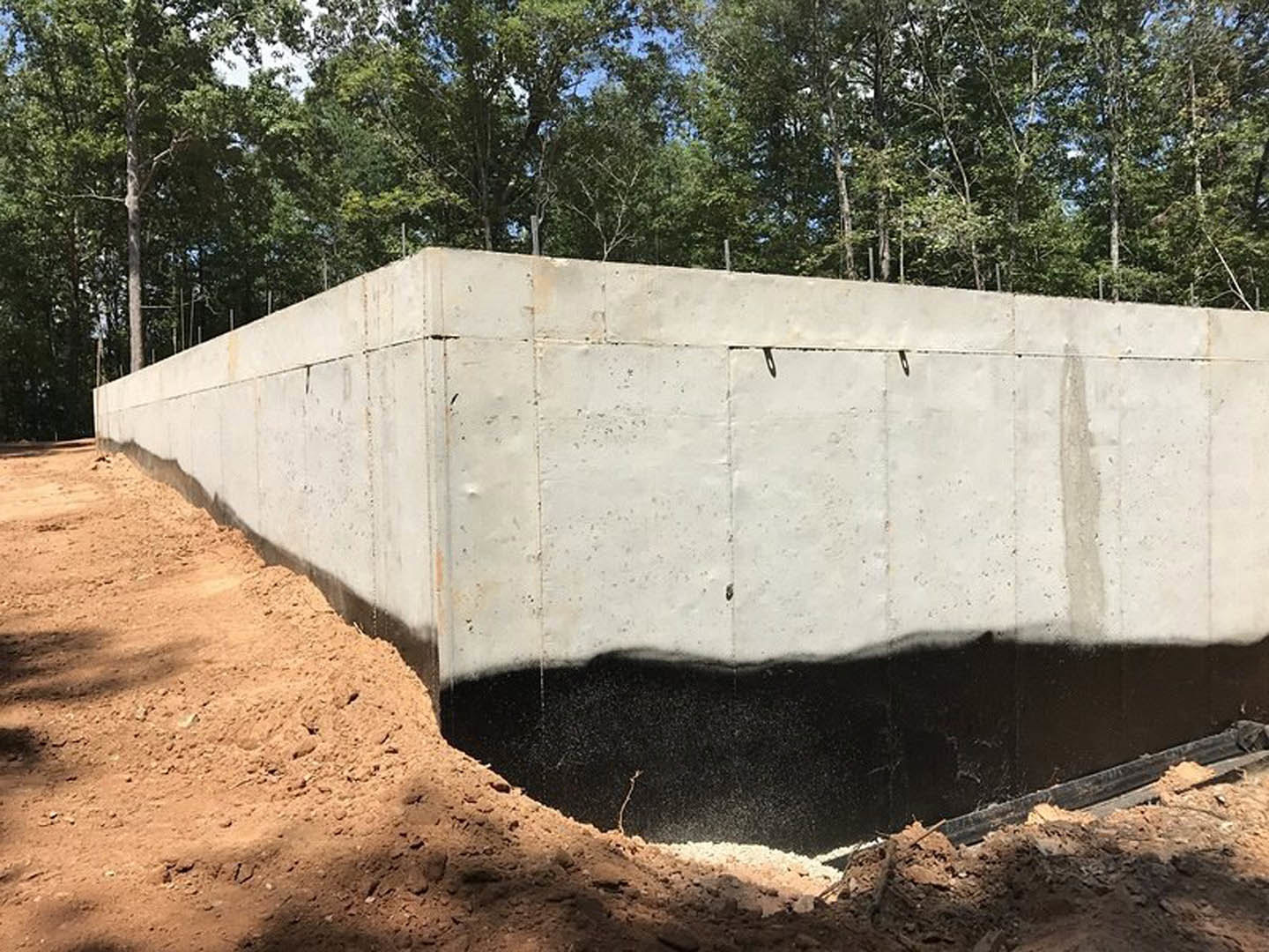 Smooth concrete foundation wall bordered by loose soil, with mature trees and sky visible in the background