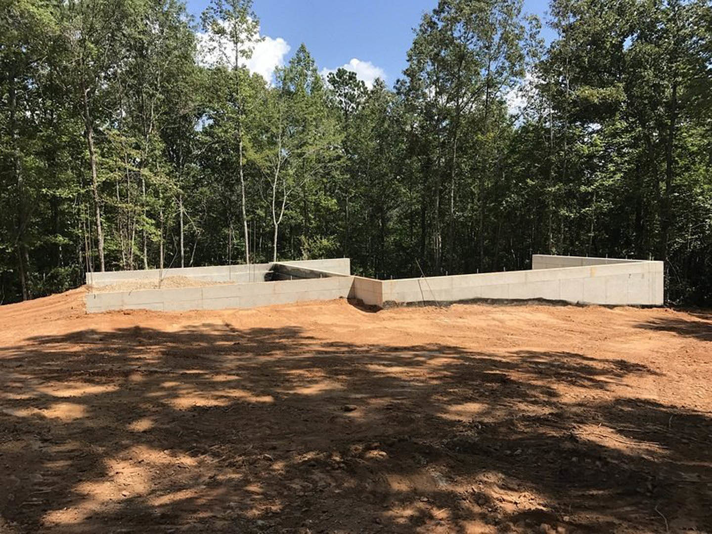 Dirt field bordered by a white concrete wall with a circular opening, white building partially visible, group of trees and blue sky with scattered clouds in the background