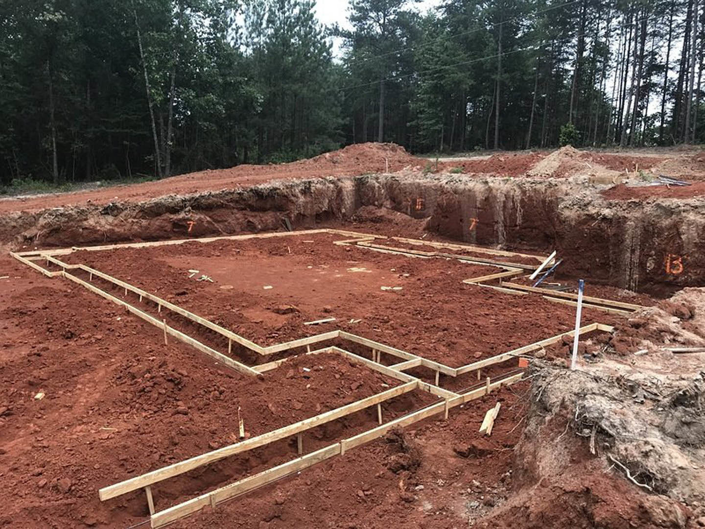 Wooden framing and beams set on a dirt foundation surrounded by trees, with a white pole and construction materials scattered across the site.