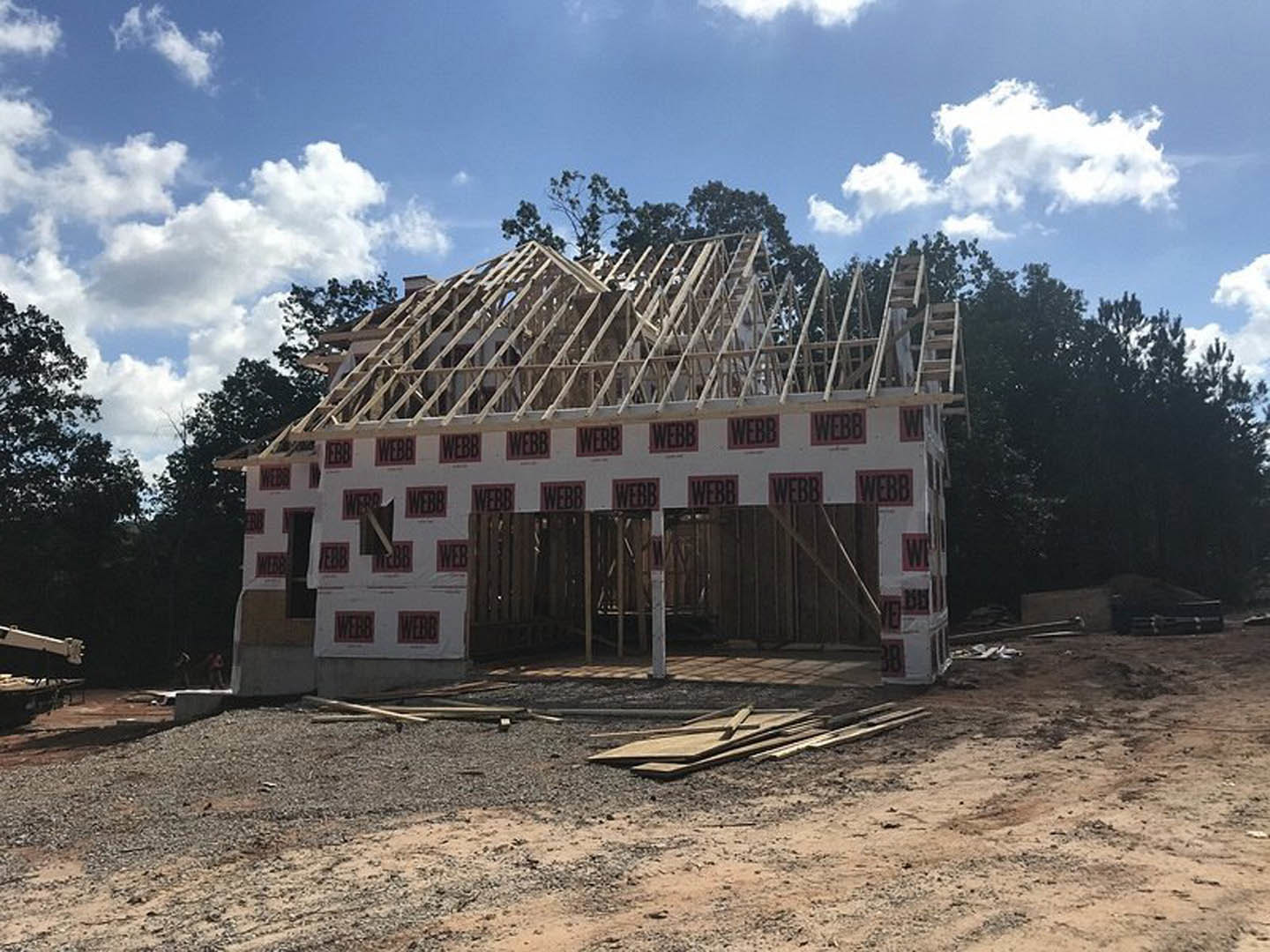Wood-framed house under construction with exposed beams, stacks of lumber on dirt ground, leafy trees and blue sky in background