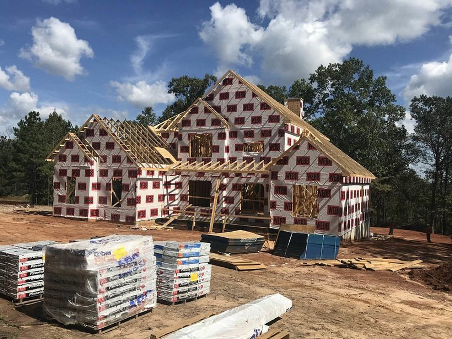 Partially built house wrapped in red and white weather barrier, stacks of pallets and cement on dirt driveway, white tarp nearby, large window installed, surrounded by trees under