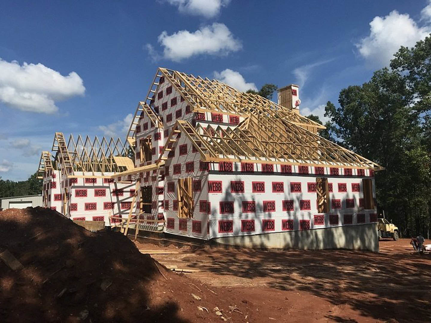 Two-story house under construction with exposed framing, red and white protective sheets, large pile of dirt in front, yellow truck parked nearby, trees and cloudy sky in